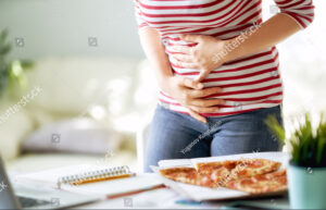 Frau isst Pizza auf dem Schreibtisch bei der Arbeit und hat Magenprobleme.